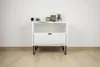 A white bedside table with a drawer and an open shelf, placed on a wooden floor against a white wall. The table has black hairpin legs and is decorated with a few items on the shelf. Podgląd zdjęcia produktu