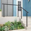 A black metal handrail installed on an outdoor staircase leading to a house entrance, with lush green plants in the foreground. Produktbild-Vorschau