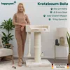 A woman stands next to a cat tree, with decorative items and plants in the background. Produktbild-Vorschau