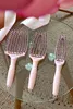 Three pink hairbrushes are arranged on a glass table with a textured rug underneath. A pink ribbon is draped across the top of the image. Produktbild-Vorschau