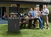 A family is gathered around a table in a backyard, with a gas grill in the foreground. Produktbild-Vorschau