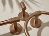 A set of copper-colored bathroom accessories, including a toilet paper holder, a towel ring, and a hook, are displayed on a textured surface. Produktbild-Vorschau
