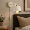 A wall lamp is mounted above a bed, next to a nightstand with a plant and books. Náhled obrázku produktu