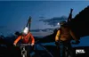 Two people on bicycles with skis on their backs, riding at dusk. Náhled obrázku produktu