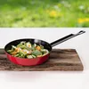 A red mini frying pan filled with green vegetables and small white flowers, placed on a rustic wooden board with a blurred green background of a meadow. Produktbild-Vorschau