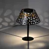 A solar-powered table lamp with a decorative metal shade is shown in a dimly lit room, casting patterned shadows on the walls and floor. Produktbild-Vorschau