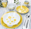 A set of yellow and white polka dot dinnerware is arranged on a light wooden surface, with some greenery and white flowers in the background. Podgląd zdjęcia produktu