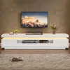 A modern white TV stand with LED lighting, featuring a large flat-screen TV mounted above it, displaying a mountain landscape. The stand holds electronic devices and decorative items. A book and glasses are placed on the rug in front of the stand. Podgląd zdjęcia produktu