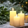 Three LED candles of varying sizes are placed on a wooden surface, with a snowy pine branch in the foreground and a blurred background of greenery and bokeh lights. Snowflakes are falling. Voir les images du produit