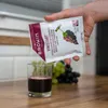 A person's hand is pouring a white powder from a "BROWIN VINISTART" packet into a glass of dark red liquid. Produktbild-Vorschau