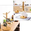 A collage of two images showing a wooden mug tree stand. The left image shows the stand on a wooden desk with two watches and a phone stand. The right image shows the stand on a kitchen counter with mugs and donuts. Produktbild-Vorschau