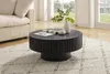 A black round coffee table with a fluted design sits on a cream-colored rug in a living room setting. A sofa is visible on the left, and windows with natural light are in the background. Produktbild-Vorschau