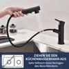 A hand is holding a black kitchen faucet spray head, with water spraying out, over a kitchen sink. The faucet base is also visible. Produktbild-Vorschau