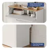 A split image showing a white shoe cabinet with a wooden top. The top image shows the cabinet with a drop-down drawer open, revealing books and decorative items. The bottom image shows the side of the cabinet, highlighting a cut-out design. Produktbild-Vorschau