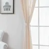 A beige curtain is tied back with a fabric tie and hangs in front of a window with white mullions. A framed picture of a building is visible on the left wall, and the arm of a white armchair is in the bottom left corner. Produktbild-Vorschau
