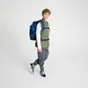 A young boy wearing a backpack and casual clothing, posing against a white background. Produktbild-Vorschau