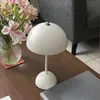 A white mushroom-shaped table lamp sits on a wooden table next to a vase of flowers and a laptop. Produktbild-Vorschau