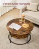 A round coffee table with a lower shelf is shown in a living room setting, with a book, a glass of water, and a pitcher on top. A sofa is visible in the background. Produktbild-Vorschau
