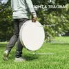 A person walks across a grassy lawn carrying a large, round, white object with a tan handle. The background is a park-like setting with trees and grass. Produktbild-Vorschau