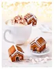 A white mug filled with whipped cream and marshmallows, next to two small gingerbread houses decorated with white icing. The background is blurred with golden bokeh lights. Produktbild-Vorschau