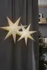 Two paper star lanterns hang in front of a dark gray curtain, with a Christmas tree and shelves visible in the background. Podgląd zdjęcia produktu