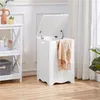 A white laundry hamper with its lid open, draped with a beige blanket, sits on a light wood floor next to a white shelf unit. The scene is set indoors with soft lighting and white curtains in the background. Produktbild-Vorschau