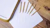 Colored pencils are scattered on a white sheet of paper, which is placed on a wooden surface. A white and gold tray is partially visible on the left side of the image. Produktbild-Vorschau
