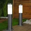 Two modern stainless steel bollard lights are illuminated in a garden setting at dusk, with a wooden fence and greenery in the background. Anteprima dell’immagine prodotto