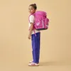 A young girl wearing a pink backpack stands against a plain background. Produktbild-Vorschau