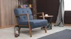 A blue armchair with wooden legs sits in a room with a bookshelf in the background. Produktbild-Vorschau