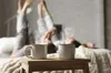 Two people lying on a bed with their legs up, holding mugs of steaming coffee. Produktbild-Vorschau