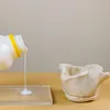 A bottle of white liquid is being poured onto a clear surface next to a small, light-colored ceramic bowl. Produktbild-Vorschau