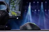 A gaming setup with a Razer mouse and headphones in front of a monitor displaying a game, with a stage-like background. Produktbild-Vorschau