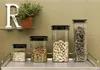 Four clear plastic food storage containers with black lids are arranged on a metal shelf against a light green wall. The containers hold different food items: oats, pasta shells, peanuts, and dried herbs. A large white letter 'R' and a potted plant are on  Anteprima dell’immagine prodotto