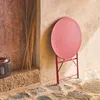 A red folding bistro table leans against a textured wall, with a lantern and dried reeds nearby. Anteprima dell’immagine prodotto