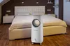 A white air cooler is placed on a wooden floor in front of a bed in a bedroom. Náhled obrázku produktu