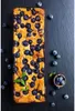 A rectangular blueberry cake is presented on a dark, slate-like surface, surrounded by scattered blueberries and a small wooden bowl filled with blueberries. Voir les images du produit