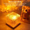 A cube-shaped lamp with a textured surface is shown on a wooden base, projecting warm, wavy light patterns onto a surface. A framed picture is visible in the background. Anteprima dell’immagine prodotto