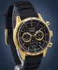 A close-up studio shot of a gold and black Seiko chronograph watch with a black rubber strap, displayed on a stand against a blue background. Náhled obrázku produktu