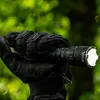A hand wearing a black tactical glove holds a black flashlight outdoors, with a blurred green background of foliage. Podgląd zdjęcia produktu