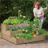 A woman watering a tiered wooden planter filled with various flowers and plants in a garden setting. Produktbild-Vorschau