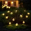 A string of solar-powered LED lights with clear, bulbous globes are draped around a green bush at night, with a blurred house in the background. Produktbild-Vorschau