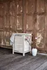 A white slatted laundry basket with a striped towel draped over the side, placed on a wooden floor in front of a rustic wooden door. A vase with pink flowers is next to the basket. Produktbild-Vorschau