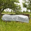 A gray tarp covers a long object resting on a stone wall, with trees and grass in the background, and rain falling. Náhled obrázku produktu