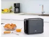 A black toaster sits on a kitchen counter next to a bowl of croissants and a small bowl of jam. A coffee maker and fruit bowl are visible in the background. Anteprima dell’immagine prodotto