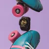 A black and gold Casio Baby-G watch is suspended between two teal roller skates with pink wheels and accents, set against a purple background. Anteprima dell’immagine prodotto