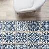 A close-up view of a patterned rug with blue and white tiles placed on a light wooden floor next to a white armchair. Produktbild-Vorschau