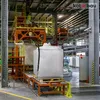 A large white industrial bag is positioned on a conveyor belt in a factory setting, with machinery and other bags visible in the background. Produktbild-Vorschau