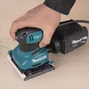 A hand is holding a Makita sander, which is resting on a wooden surface. A dust bag is attached to the sander. Anteprima dell’immagine prodotto