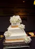 A square, multi-tiered wedding cake decorated with white flowers and gold polka dots, displayed on a silver cake stand. Náhled obrázku produktu